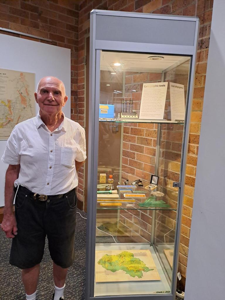Former cartographer Warren Hardy standing next to display of personal collection at Exhibition opening 4 November 2024.